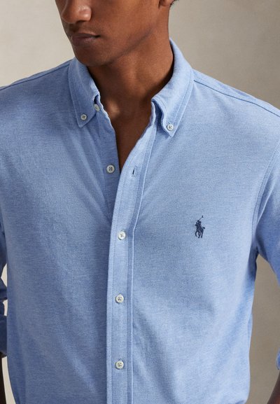 Γαλάζιο πουκάμισο με κουμπιά από υφή υφάσματος, με κλασικό γιακά και κεντητό λογότυπο στην αριστερή πλευρά του στήθους.