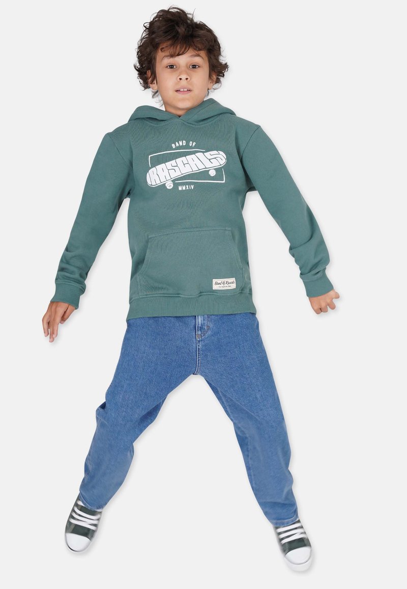 Grüne Kapuzenjacke mit weißem Grafikdesign und Kängurutasche, kombiniert mit lockeren blauen Jeans und grünen Turnschuhen mit weißen Streifen.