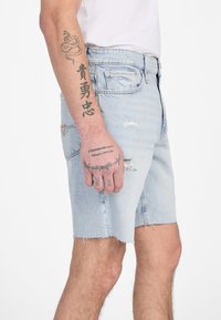 Shorts en jean bleu clair avec des bords effilochés, cinq poches et un délavage léger. Portés avec une chemise blanche, avec des tatouages visibles sur le bras.