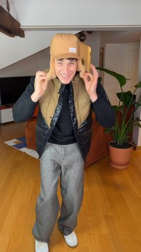 Casaco acolchoado castanho com pormenores pretos, chapéu castanho combinando com abas para as orelhas, calças cinzentas texturizadas e sapatilhas brancas, num ambiente doméstico.