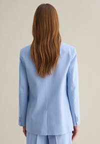 Blazer su misura azzurro chiaro con maniche lunghe, spacco posteriore e polsini con bottoni, realizzato in tessuto liscio. Indossato sopra pantaloni abbinati.