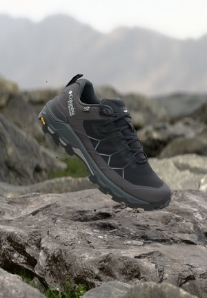 Czarno-szary but do wędrówek Columbia Titanium w powietrzu nad skalistym terenem z górskim tłem, ukazujący wytrzymałą podeszwę i outdoorowy design.