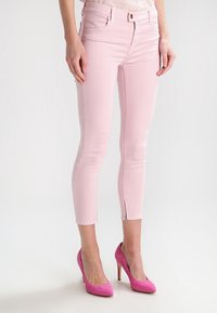 Mujer vestida con pantalones recortados ajustados de color rosa claro con cremalleras en los tobillos y zapatos de tacón alto de color rosa brillante, de pie sobre un suelo blanco.