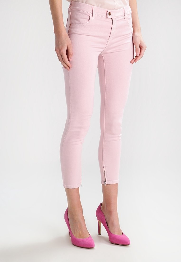 Mujer vestida con pantalones recortados ajustados de color rosa claro con cremalleras en los tobillos y zapatos de tacón alto de color rosa brillante, de pie sobre un suelo blanco.