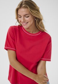 T-shirt rouge en tissu lisse, avec des manches courtes, un col rond, et un passepoil blanc le long du col et des manches.