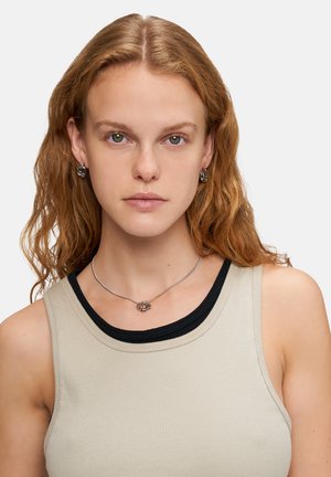 Silberne Halskette mit einem floralen Anhängerdesign und passenden silbernen Ohrsteckern. Das Model trägt ein hellbeiges, geripptes Tanktop mit schwarzem Saum.