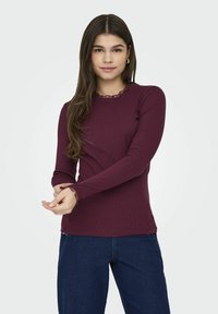 Maglietta a maniche lunghe di colore bordeaux realizzata in tessuto a costine, con scollatura e polsini a ruches, abbinata a pantaloni in denim blu.