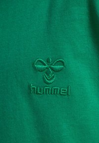 Zielony materiał z wyhaftowanym logo marki Hummel, przedstawiającym stylizowaną pszczołę nad nazwą marki.