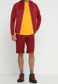 Chaqueta con capucha roja y cremallera sobre una camiseta amarilla con un gráfico de montaña, combinada con pantalones cortos rojos y botas de senderismo negras con acentos rojos.