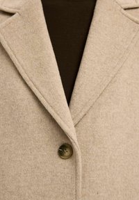 Beige wollen mix jas met een geknopt kraag, enkele knoopsluiting en een zachte textuur, gedragen over een donkerbruine top.