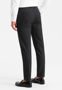 Pantalons de costume noirs fuselés avec une texture lisse, comportant deux poches arrière, une fermeture à bouton et des coutures détaillées. Portés avec des mocassins noirs.