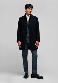 Cappotto blu scuro con colletto alla coreana e chiusura con bottoni a un petto, indossato sopra un completo grigio a quadri e scarpe eleganti nere.