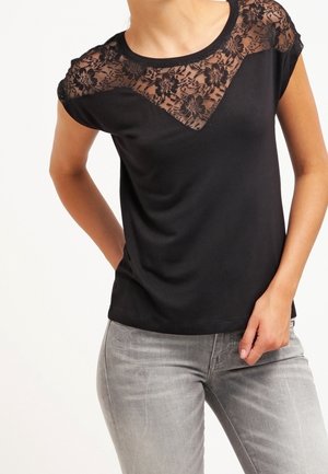 Frau trägt ein schwarzes Kurzarm-Top mit floraler Spitze über Schultern und Brust, kombiniert mit hellgrauen Jeans.
