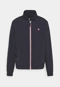 Męska granatowa kurtka przeciwwiatrakowa z wysokim kołnierzem, zapięciem na przedni zamek błyskawiczny, elastycznymi mankietami oraz małym czerwono-białym logo na piersi.