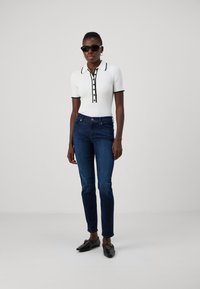 Witte geribbelde poloshirt met zwarte details en knopen, gecombineerd met donkerblauwe jeans en zwarte instappers. Model draagt een zonnebril.