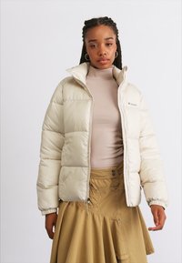 Jachetă puffer de culoare bej deschis, cu fermoar în față, guler înalt și manșete elastice. Purtată peste un pulover cu guler înalt de culoare c�ăpru deschis și o fustă bej, are logo-ul Columbia.