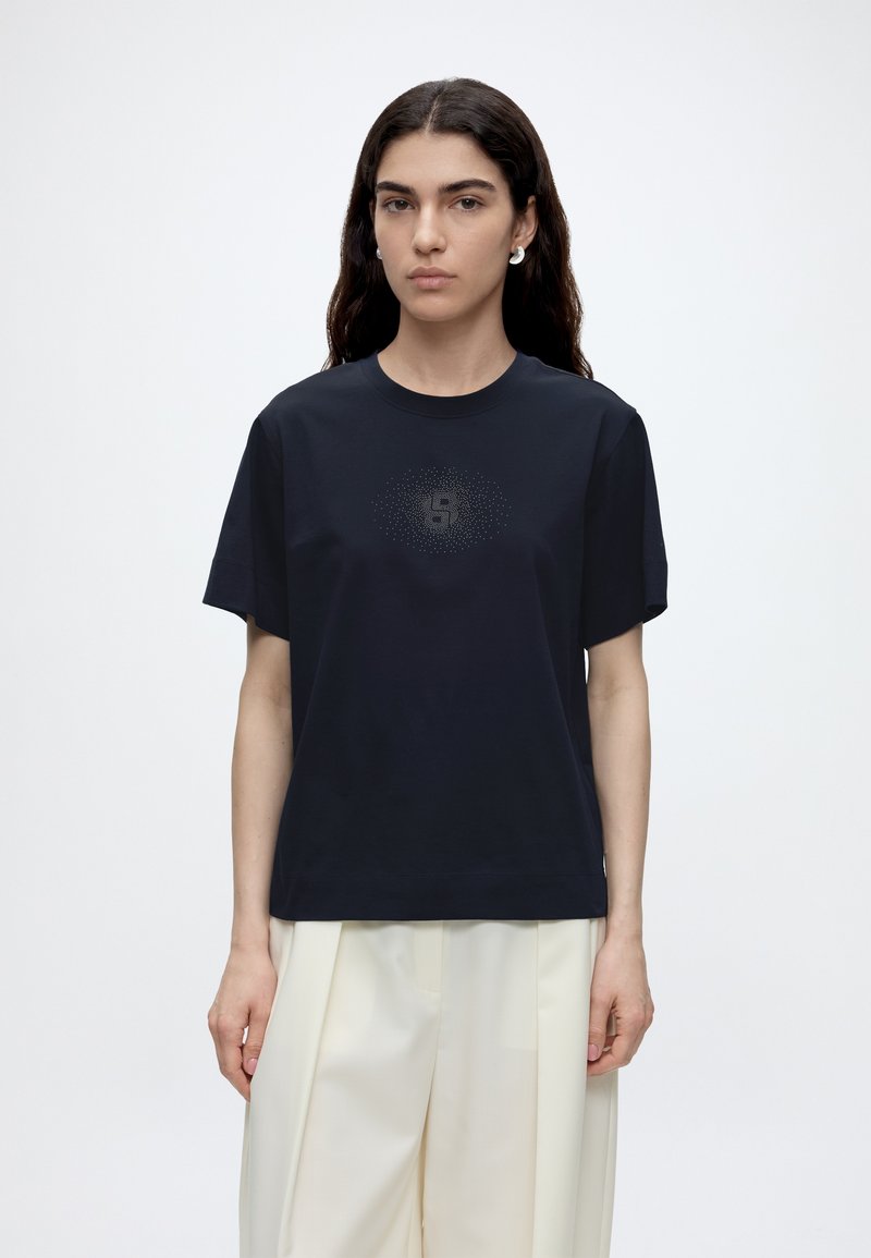 Femme aux longs cheveux foncés portant un T-shirt noir à manches courtes avec un motif délicat à pois et un pantalon large couleur crème clair.