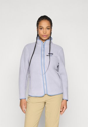 Vrouw met gevlochten haar die een lichtpaarse fleece jas met blauwe trim en beige broek draagt, staande tegen een effen achtergrond.
