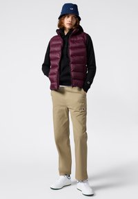 Gilet imbottito bordeaux sopra una felpa nera con cappuccio, pantaloni beige e sneakers bianche. Cappello a secchiello blu con dettaglio del logo. Tessuto morbido, vestibilità casual.