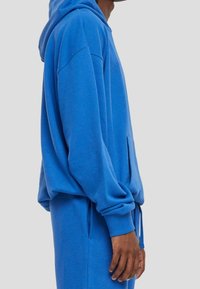 Sudadera azul de tela suave con una capucha amplia, bolsillo tipo canguro y cordones en el dobladillo. Presenta un corte holgado y puños de canalé.