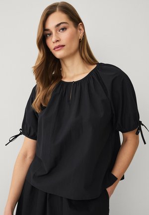 Vrouw met lang bruin haar die een zwarte losse korte mouwen blouse draagt met strikjes op de mouwen, staand tegen een lichtgrijze achtergrond.