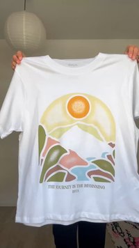 Άσπρο T-shirt που κρατιέται ψηλά, με στυλιζαρισμένο ήλιο, βουνά, ποτάμι και κείμενο "ΤΟ ΤΑΞΙΔΙ ΕΙΝΑΙ Η ΑΡΧΗ" με το όνομα της μάρκας RVCA.