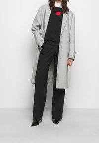 Abrigo largo gris claro con un patrón texturizado, sudadera negra con cuello redondo, pantalones anchos negros y tacones negros puntiagudos. Acento de logo rojo en la sudadera.