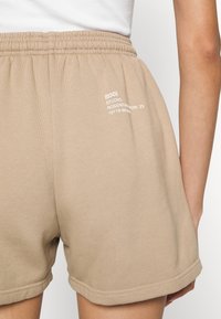 Shorts beiges à taille élastique avec texte imprimé blanc sur le côté droit, portés avec un haut blanc, visibles de la taille jusqu'au milieu des cuisses.