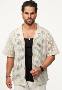 Gestricktes Kurzarmhemd in Creme und Hellblau, mit einem durchbrochenen Muster. Vorgeknöpfte Vorderseite und umgeschlagener Kragen. Schwarzes Top darunter.