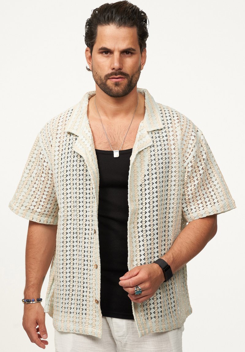 Gestricktes Kurzarmhemd in Creme und Hellblau, mit einem durchbrochenen Muster. Vorgeknöpfte Vorderseite und umgeschlagener Kragen. Schwarzes Top darunter.