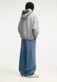 Harmaa huppari isolla hupulla ja mustalla logolla takana, yhdistettynä väljälahkeisiin sinisiin farkkuihin ja valkoisiin lenkkareihin.