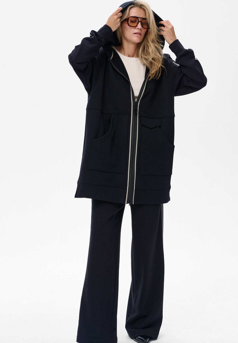 Czarna bluza z kapturem na zamek błyskawiczny z dużymi kieszeniami, w połączeniu z czarnymi spodniami typu wide-leg; oba elementy wykonane z miękkiego materiału z minimalnymi detalami.