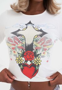 Nahaufnahme eines Oberkörpers, der ein weißes Langarmshirt mit einem Design aus einem Kreuz, einem roten Herzen, Rosen, gelben Blumen und Schmetterlingen trägt.