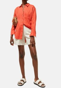 Locker sitzendes oranges Leinenhemd mit langen Ärmeln, Knopfleiste vorne, einzelne Brusttasche, kombiniert mit hellen Shorts und weißen Sandalen.