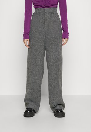 Personne portant un pantalon gris taille haute à jambes larges, des chaussures à plateforme noires et un haut violet à manches longues froncé, debout devant un fond blanc.