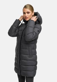 Woman adjusting hood of long, dark gray quilted winter coat with front zipper and side pockets, standing against white background.