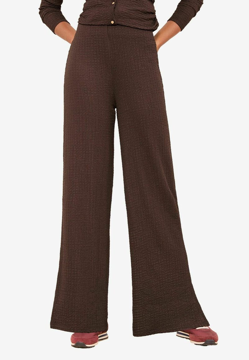 Barna, széles szárú nadrág texturált anyagból, magas derékkal és zsebekkel. Piros, hétköznapi tornacipővel párosítva.