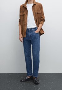 Chemise en daim marron, T-shirt blanc, jean en denim bleu et mocassins noirs. La chemise est dot ée de deux poches poitrine et d'une coupe décontractée.