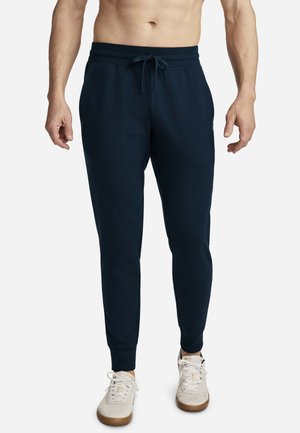 Mann, der dunkelblaue Jogginghosen mit Kordelzugbund und weißen Sneakers trägt, vor einem schlichten hellen Hintergrund stehend.