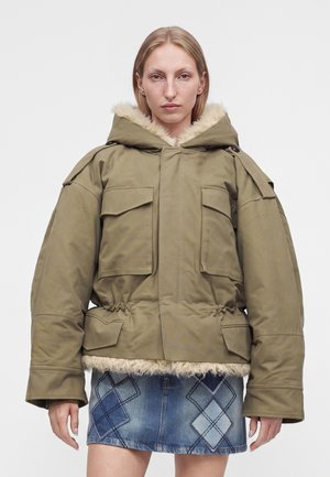Mulher vestindo casaco oversized kaki com forro de pelúcia e bolsos aplicados, combinado com uma saia de ganga azul com padrões argyle.