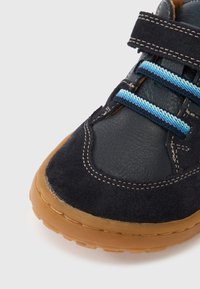 Σκούρο μπλε παπούτσι από σουέντ με υφασμάτινη υφή, διαθέτει ανοιχτό μπλε κορδόνια και σόλα από καουτσούκ τύπου γκαμ. Λουράκι με βέλκρο για ρυθμιζόμενη εφαρμογή.