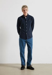 Chemise bleu marine à boutons avec un petit logo sur la poitrine, associée à un jean bleu et des chaussures noires. La chemise a un col classique et des manches longues.