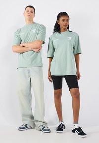 Divi modeļi stāv plecu pie pleca. Vīrietis valkā gaiši zaļu oversized T-kreklu, izbalinātas džinsus un tumšas sporta kurpes. Sieviete valkā atbilstošu T-kreklu, melnas šortus un melnas sporta kurpes.