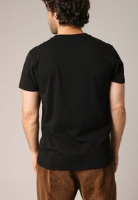 T-shirt noir en coton à manches courtes, col rond, avec un design uni. Porté avec un pantalon en velours côtelé marron, montrant la vue arrière.