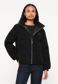 Czarna kurtka puchowa z teksturą w romby, zapięcie na zamek, wysoki kołnierz i elastyczne mankiety. Nosi się ją na czarnym swetrze i dżinsach.