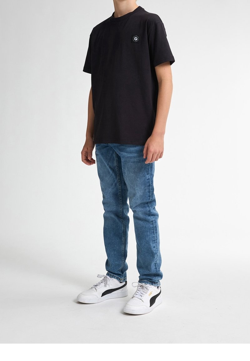 Personne portant un t-shirt noir, un jean bleu et des baskets blanches, debout devant un fond blanc uni.