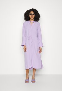 Vestido longo lilás de mangas compridas com cintura elástica, decote em forma de buraco de chave e punhos amplos. Combinado com sandálias lilás de salto robusto e óculos de sol.