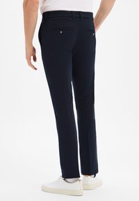 Granatowe spodnie o dopasowanym kroju, z dwiema tylnymi kieszeniami zapinanymi na guziki oraz gładką fakturą. W zestawie z białymi sneakersami.