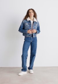 Chaqueta de mezclilla con un cuello de piel blanca, jeans de mezclilla azules y zapatillas blancas. Presenta un diseño clásico con cierres de botones y bolsillos.