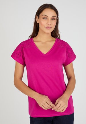 Femme aux longs cheveux foncés portant un t-shirt rose vif à col en V avec des manches courtes brodées, debout devant un fond gris clair.